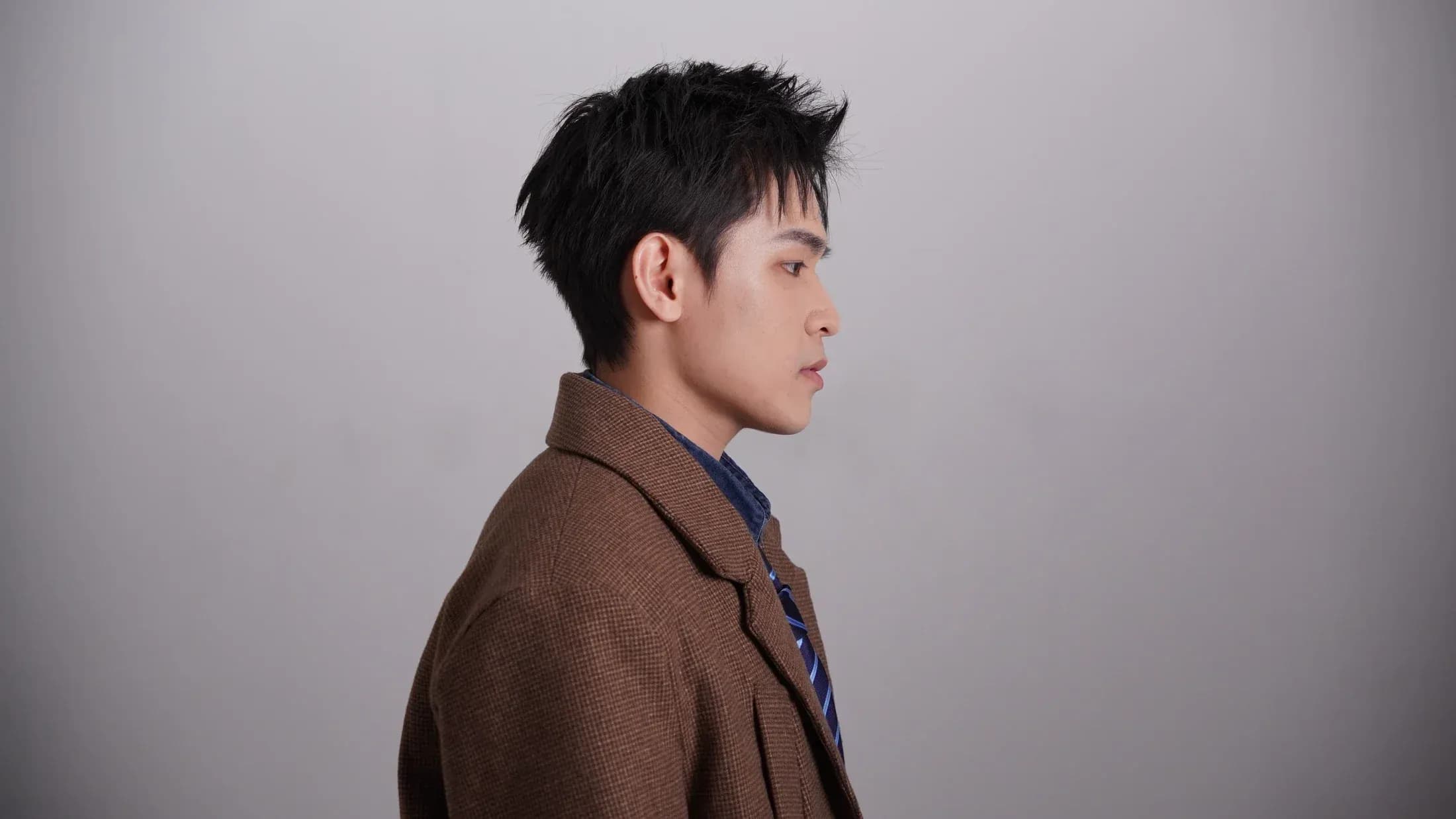 KIDEN in profile wearing a brown jacket against a grey studio background